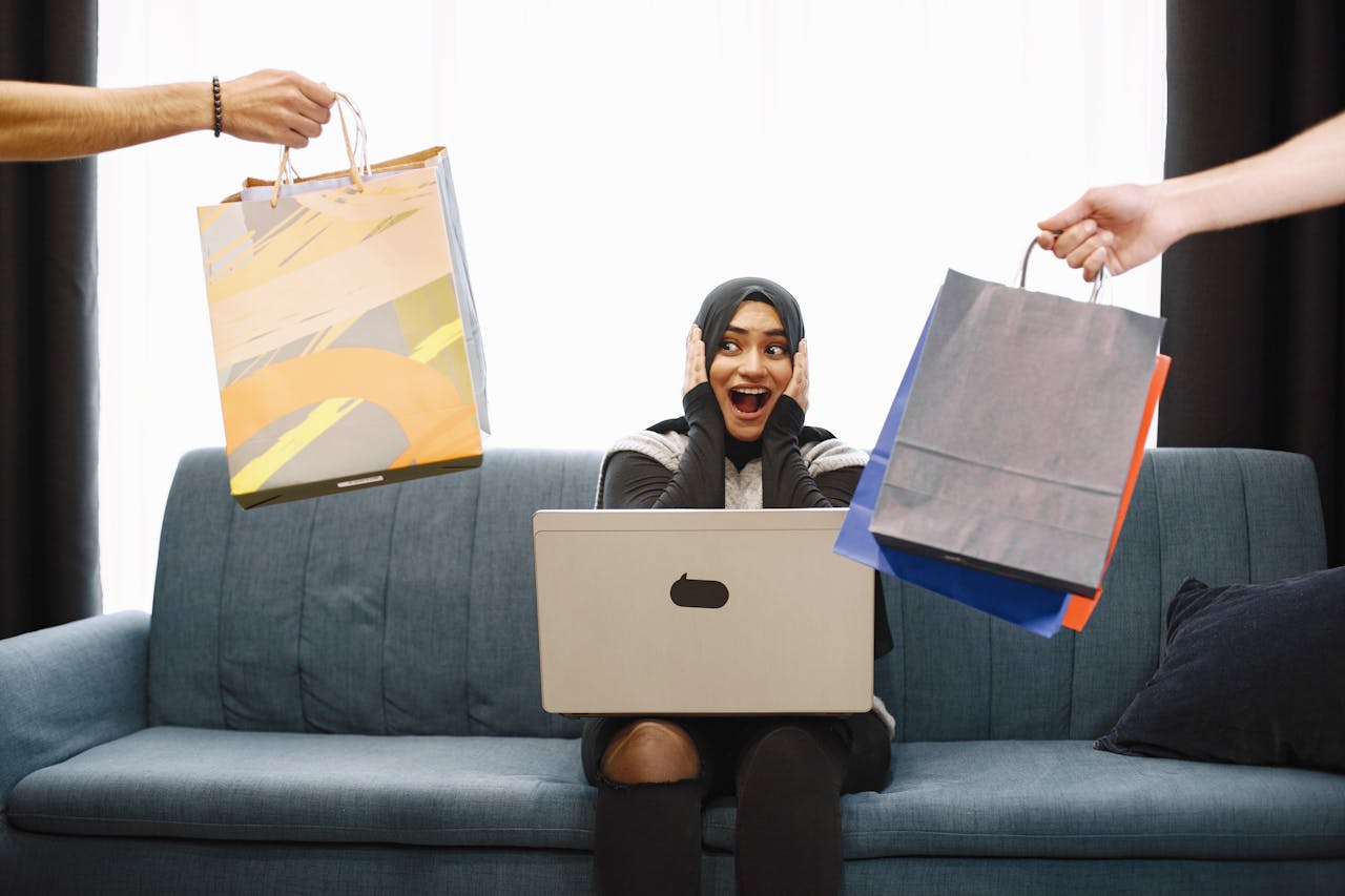 services-02 A woman in a hijab excitedly shopping online, receiving bags while sitting on a couch with a laptop.